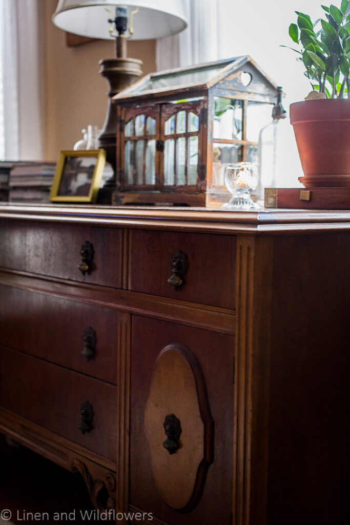 5 Uses for an Antique Sideboard- A beautiful antique side in front of a window in the livingroom styled with stacks of books, a gold landscpae frame, oil lamp, terraium with houseplants, candle, vintage spritzer & another house plant resting on an old cigar box.