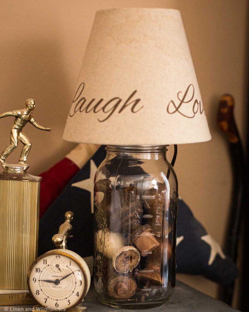 diy mason jar lamp-mason jar lamp filled with antque finials on a small blue bed table with a antique metal clock, bowling trophy & a folded American flag.