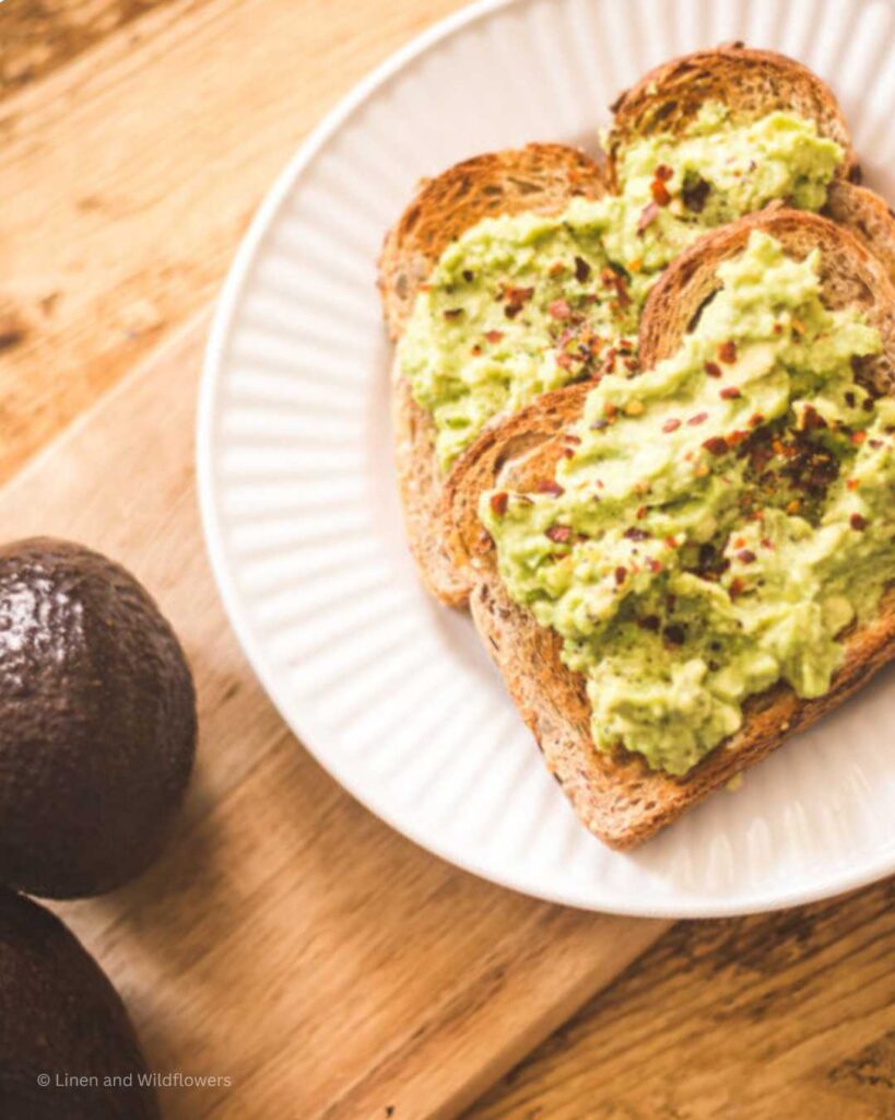 A white plate with two slices of toasted bread with avocado spread. Next to it are two fresh avocados.