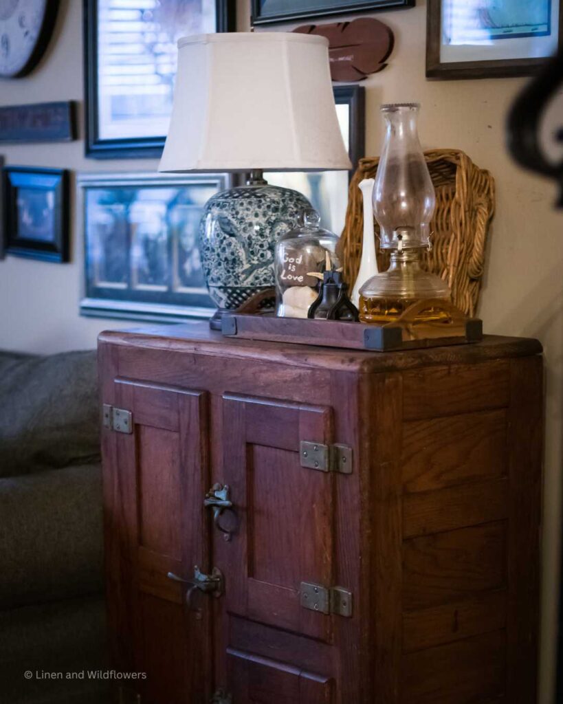 A antique icebox with a oriental lamp, cloche willed with seashells & a rock that says "God is Love" along with a liberty bell & an oil lamp. Behind it is a square basket, above is a wall gallery of photos & memorabilia.