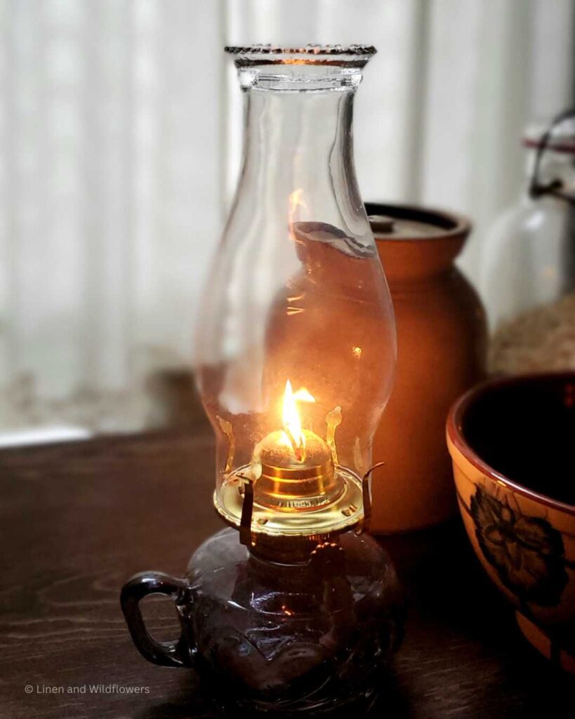 a lit oil lamp on a table with a crock, bowl & large mason jar filled with dry goods.