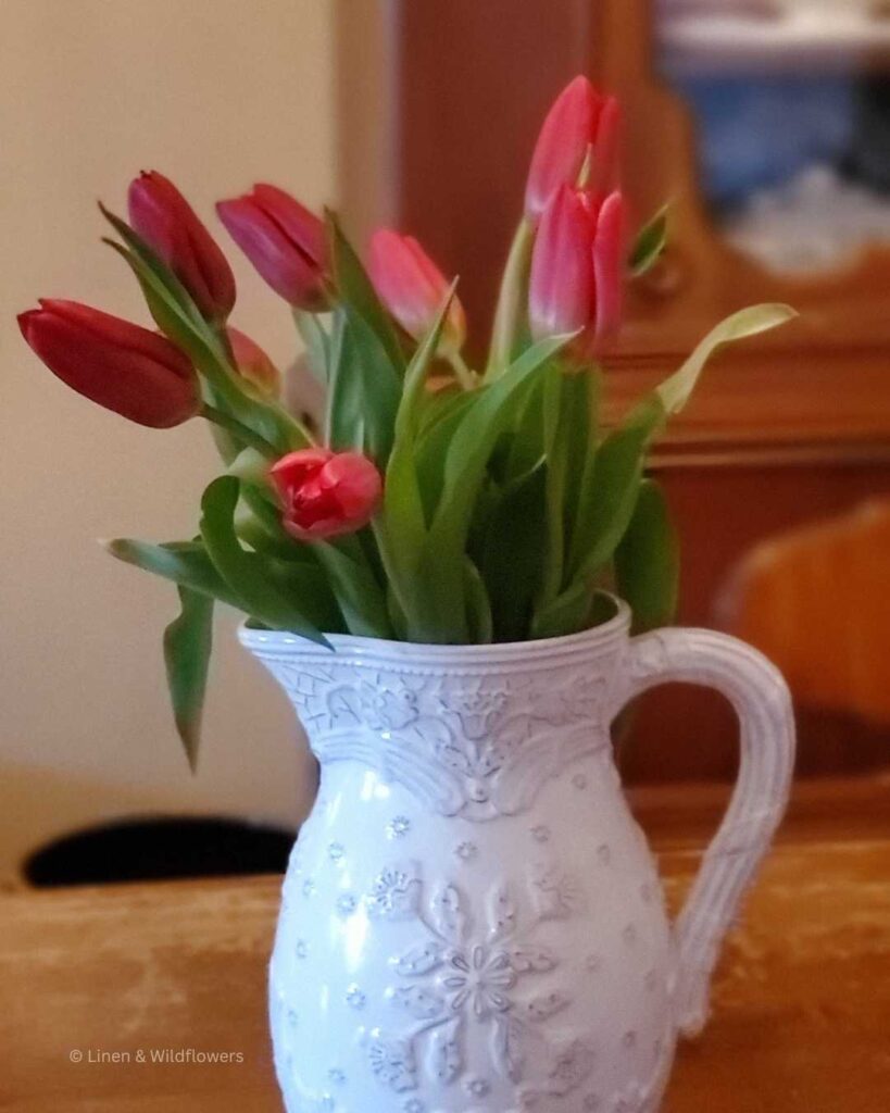A white pitcher with a bouquet of Red Tulips on a dining table in front of a china cabinet.