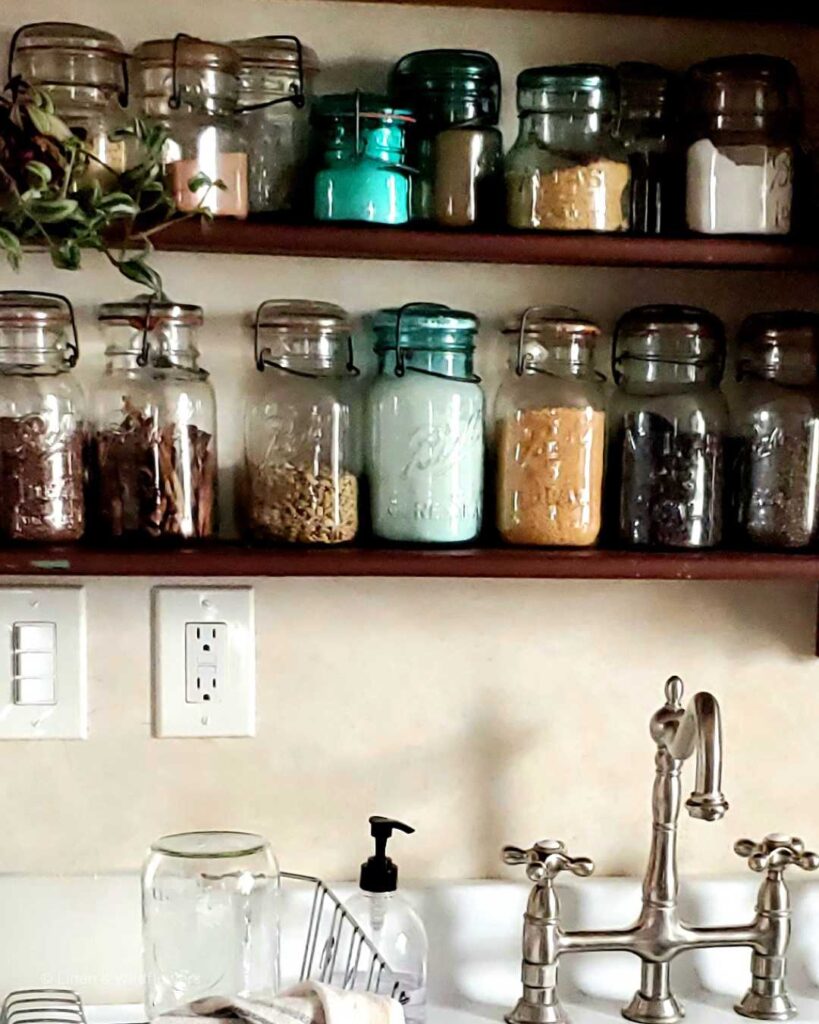 A two tiered wall shelf above the vintage sink with a large assortment of small & large vintage mason jars filled with dry goods & a wondering jew plant on the top shelf.
