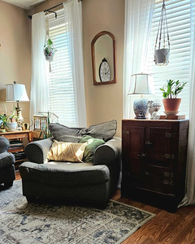 A cozy living room with a Victorian ice box & in the corner is a end table with plants, lamp & a stack of decor books.