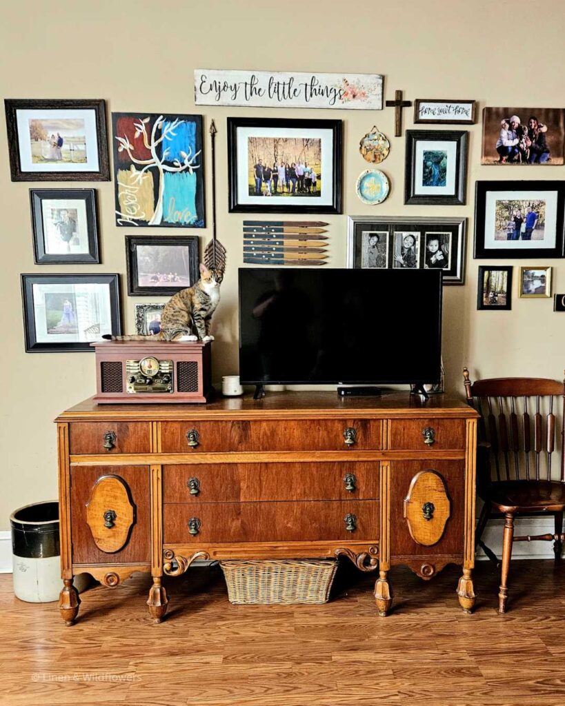 A large gallery wall, A victorian side board used as a tv stand & a vintage style radio with a Calico cat sitting on top of it. Next to the sideboard is a wooden chair & on the other end is a antique brown & white crock,