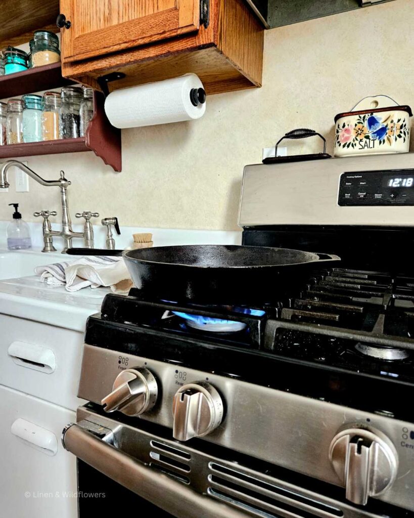  A large cast iron skillet drying on a dirty lit stove next to a vintage sink.