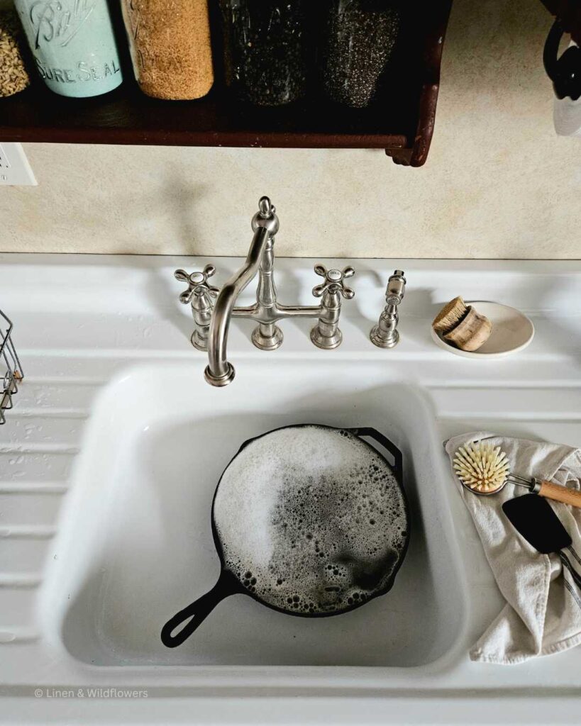A large cast iron skillet soaking in hot soapy water in a vintage sink. on the drainboard is a ironstone dish with a wooden scrubber, a long handle scrub brush & scraper on a tea towel.