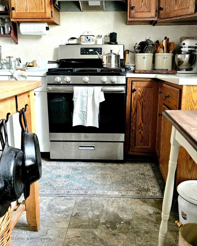A working kitchen with a stainless steel stove, vintage sink, butcher block with cast iron hanging from it, cast iron skillets stacked on the stove, crocks filled with utensils & a kitchen aid on the counter.