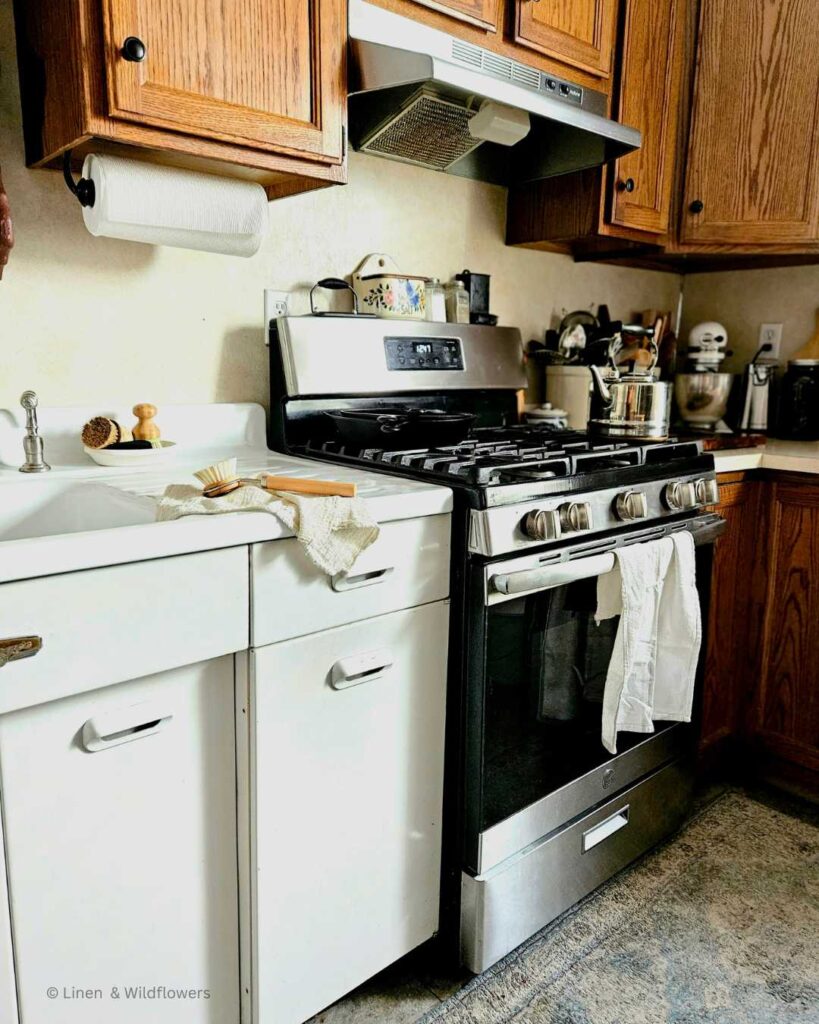 A working kitchen with a stainless steel stove, vintage sink, butcher block with cast iron hanging from it, cast iron skillets stacked on the stove, crocks filled with utensils & a kitchen aid on the counter.
