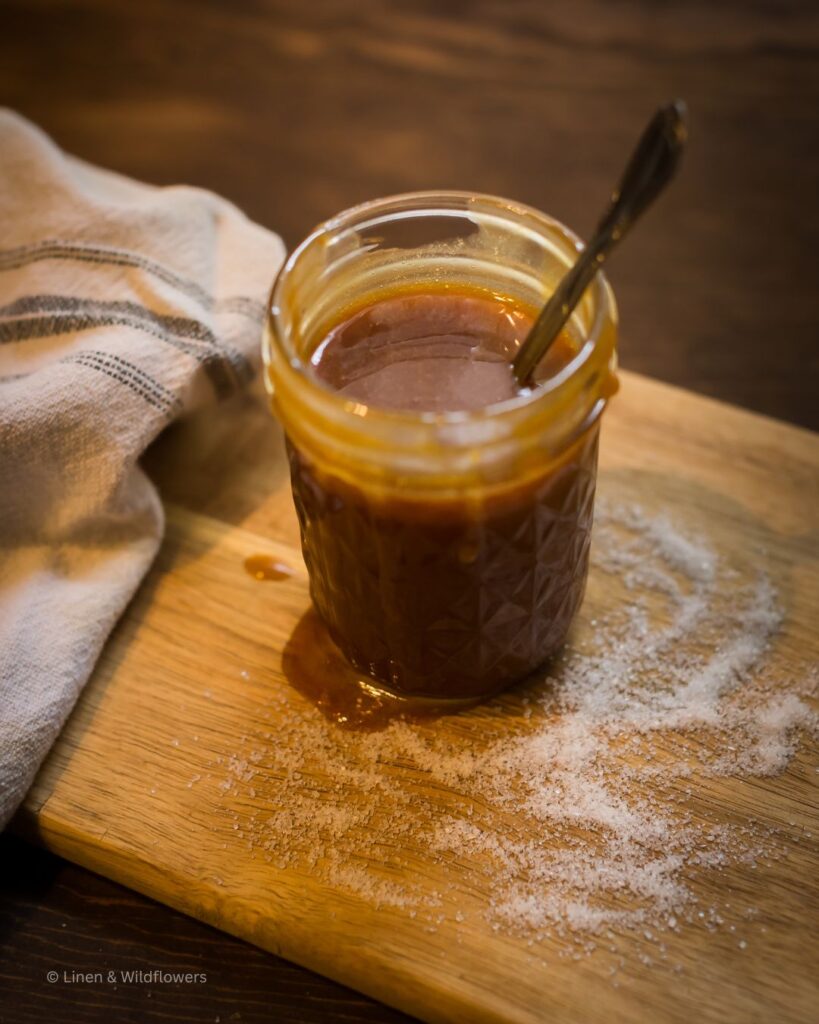 A tea towel next to a mason jar with rich amber color caramel sauce spilled over the jar onto a cutting board with sprinkles of sea salt with a serving spoon.