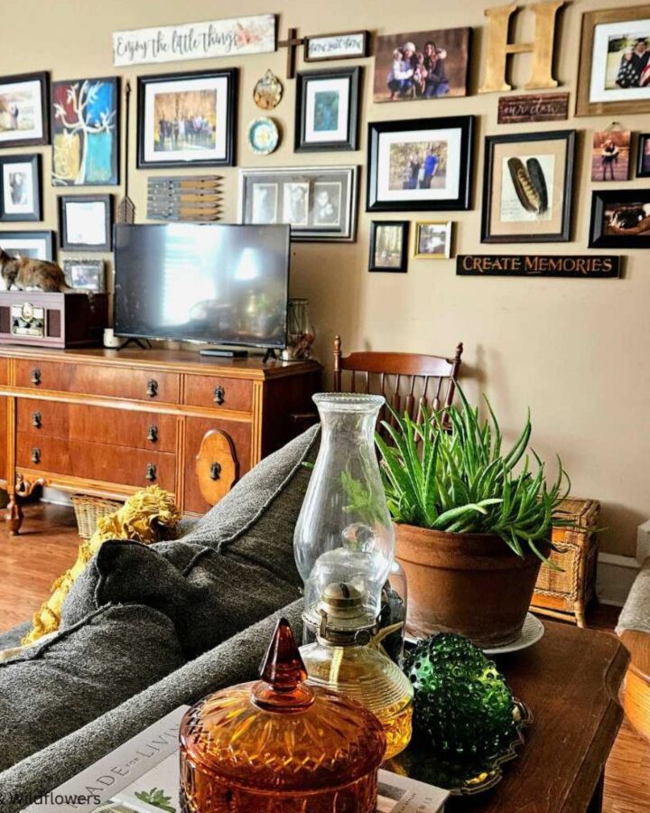 A antique sideboard in a cozy living room used to hold a tv & vintage stereo. Behind a the sofa is a sidetable with a aloe vera plant, oil lamp, amber color candy dish & a fenton circle dish.