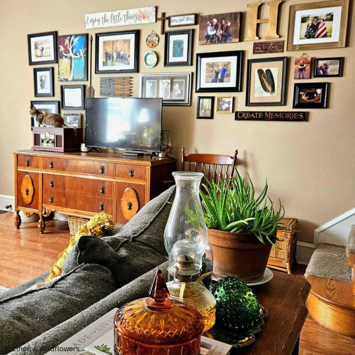 A antique sideboard in a cozy living room used to hold a tv & vintage stereo. Behind a the sofa is a sidetable with a aloe vera plant, oil lamp, amber color candy dish & a fenton circle dish.
