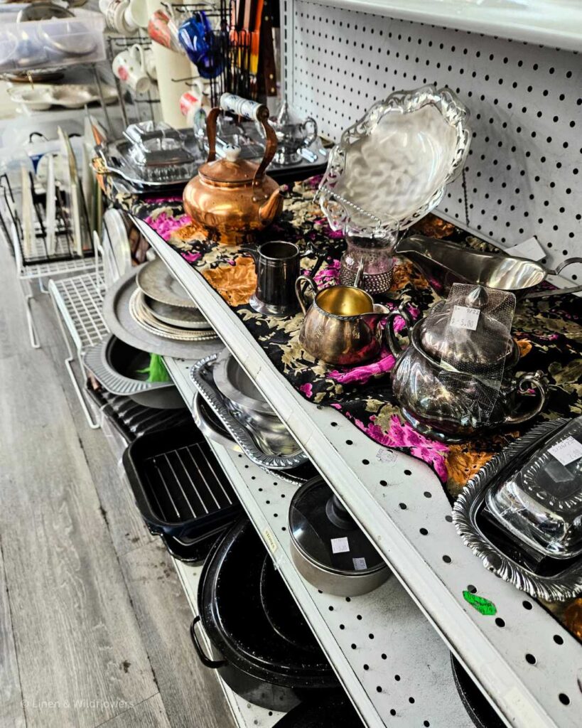 Shelves in a Goodwill Thrift Store filled with platter, silver & brass dinnerware & baking trats & roasting pans.