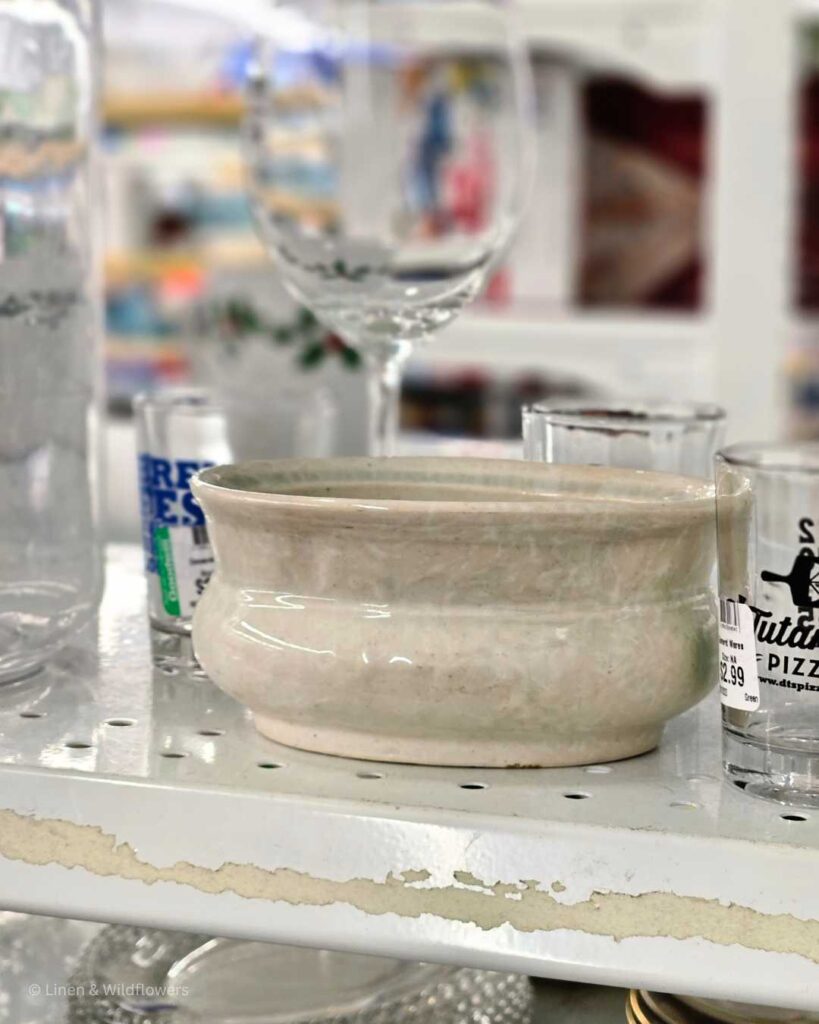 A shelf filled with a variety of glassware & a cute little crock on a shelf in a thrift store.