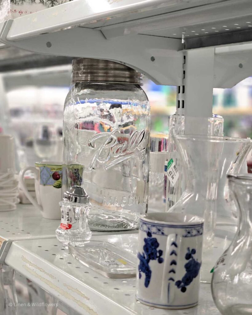 A shelf filled with a mugs, glassware & a very large 2-gallon Bal Mason Jar.