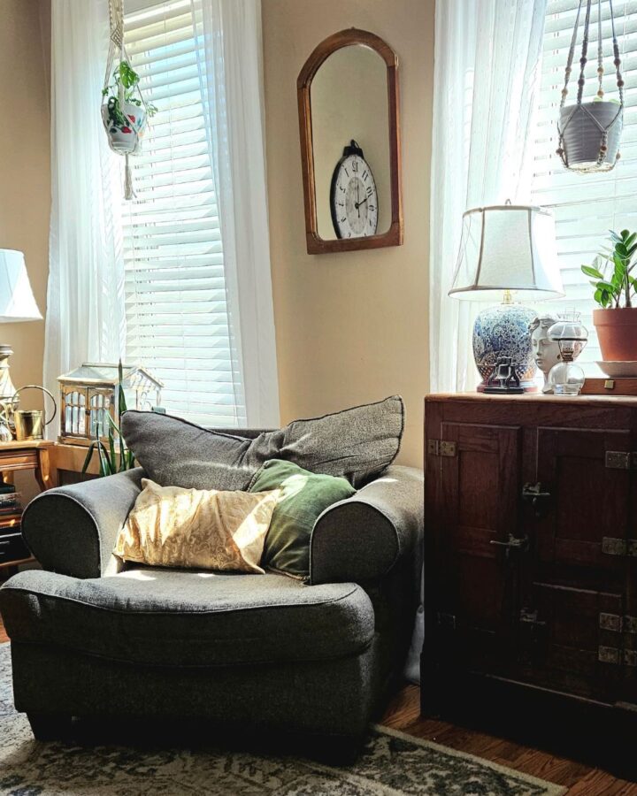 A cozy chair in a living room next to an antique icebox with a Zz Plant & oriental lamp on top. Hanging at the windows are sheer curtains & plants. Next to the couch is an antique tv stand used as an end table with old books, a lamp & house plants to liven up the space.