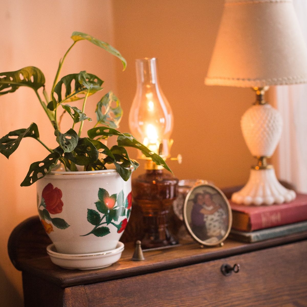 A secretary desk in the corner of a room with home decor such as a monstera plant, lit oil lamp with a amber base, a photo of Dusty with her beloved grandmother & a milkglass lamp sitting on two old books.