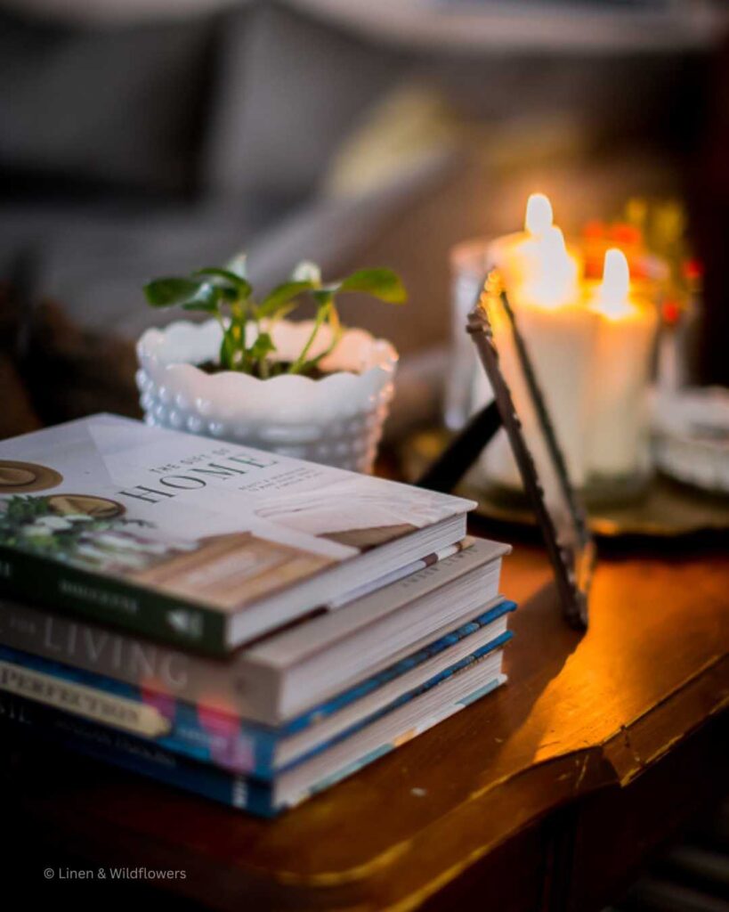 A console table with a stack of home decor books, potho plant in a milk glass planter, a framed photo & lit candles.