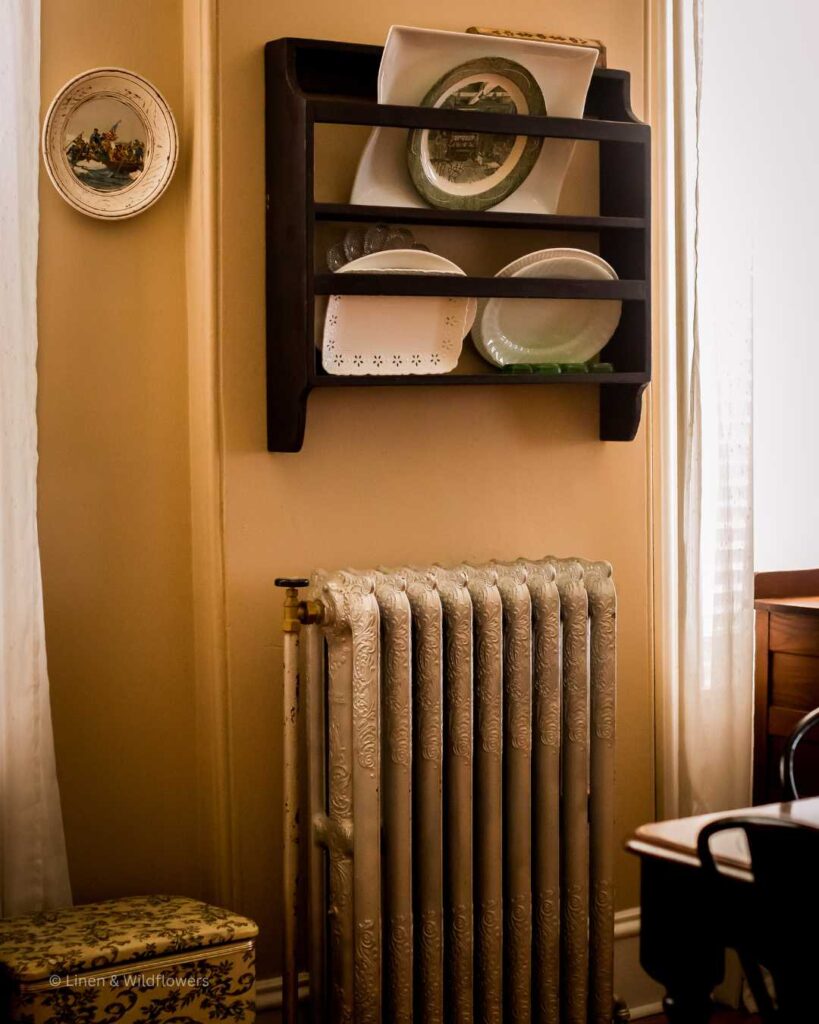 A diningroom decorated with a palte rack on the wall filled with decorative platteres, next to it is a handmade plate representing "Washington crossing the Delaware Emanuel Leutze', under the plate rack is the original cast iron radiator form a 1885 house. Next to the radiator is a vintage floral sewing box.