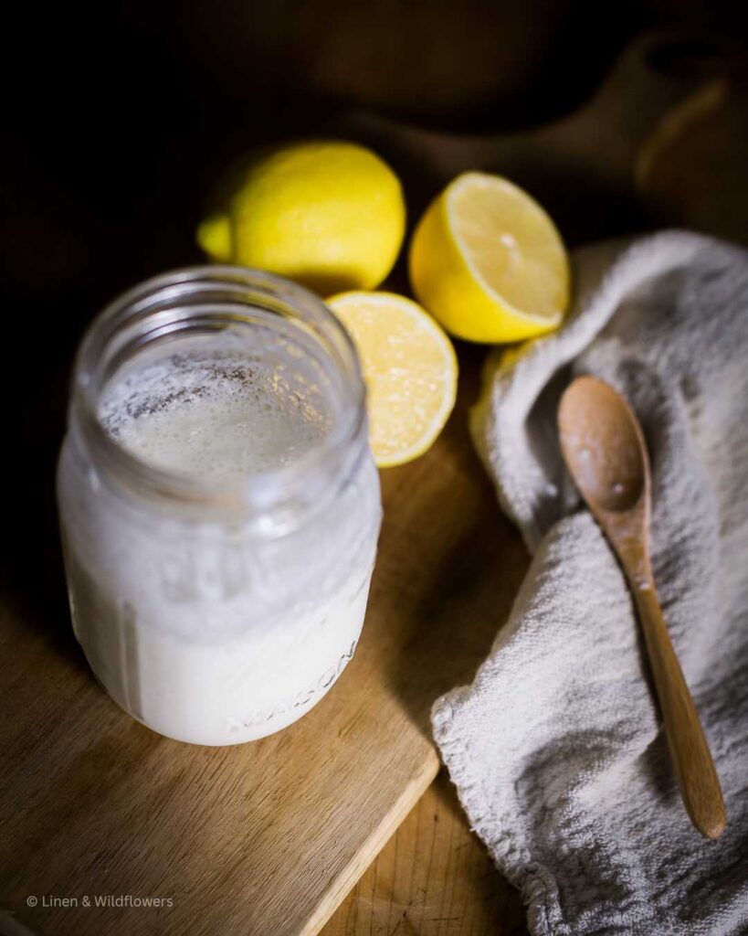 On a cutting board is a mason jar with homemade buttermilk using only 2 ingredients, a wooden spoon on a tea towel & lemons.