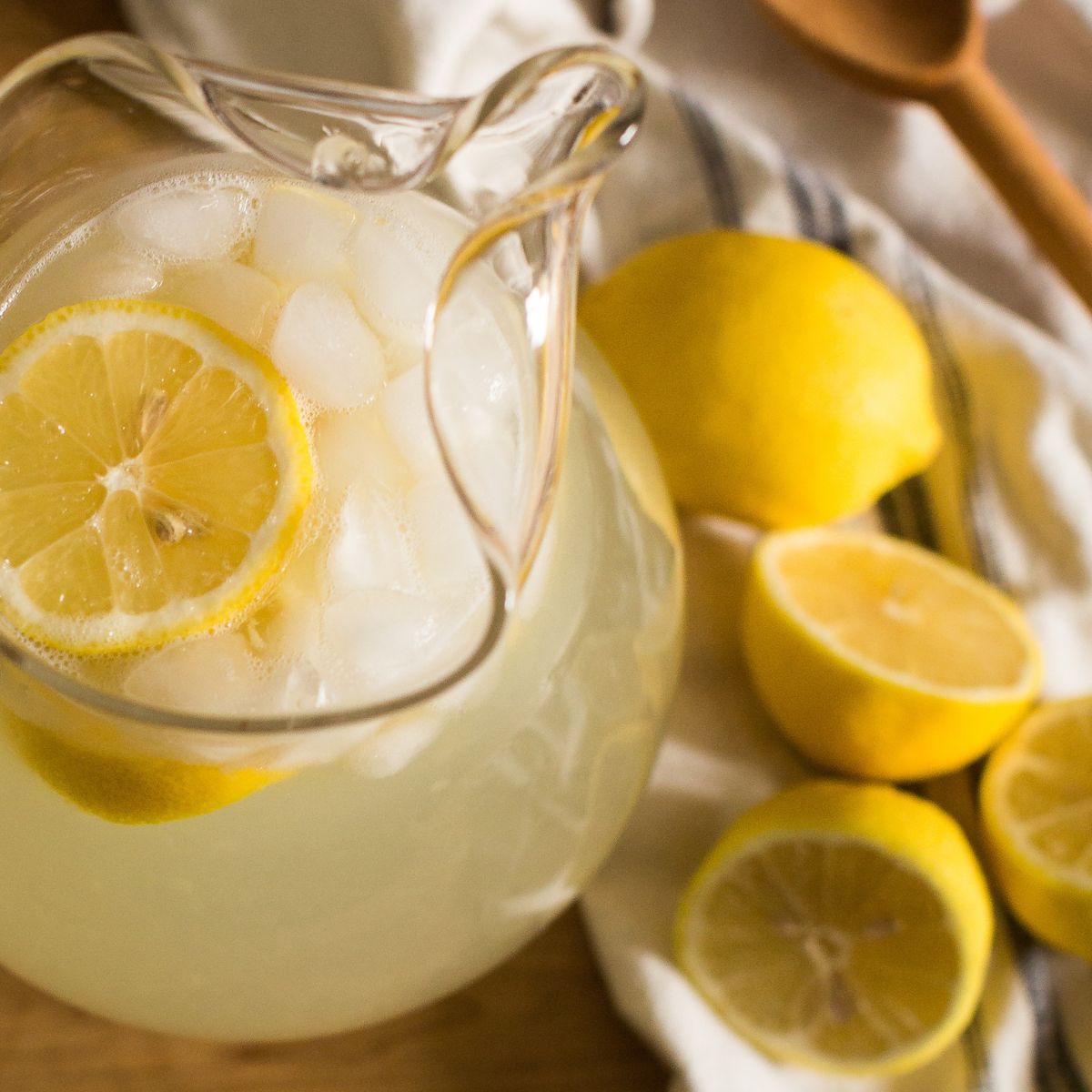 A pitcher of delicious & refreshing homemade southern sweet lemonade. Next to the pitcher is a tea towel with a wooden spoon stirrer & juicy lemons.
