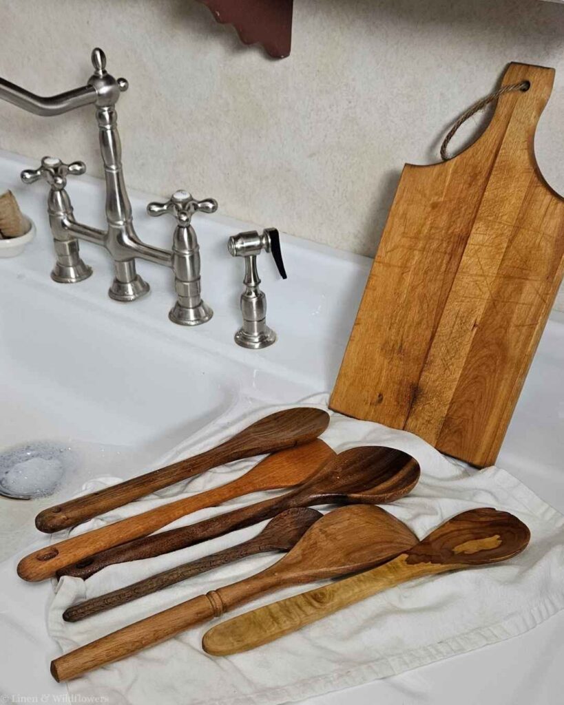 Wooden spoons drying on a linen towel beside a wood cutting board at a farmhouse sink.