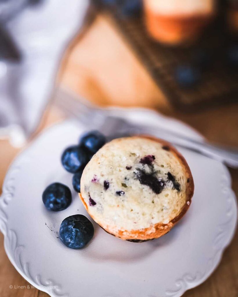 a blueberry muffin fresh out of the oven with a side of fresh blueberries on an ironstone dish with a fork ready to devour in the burst of deliciousness.