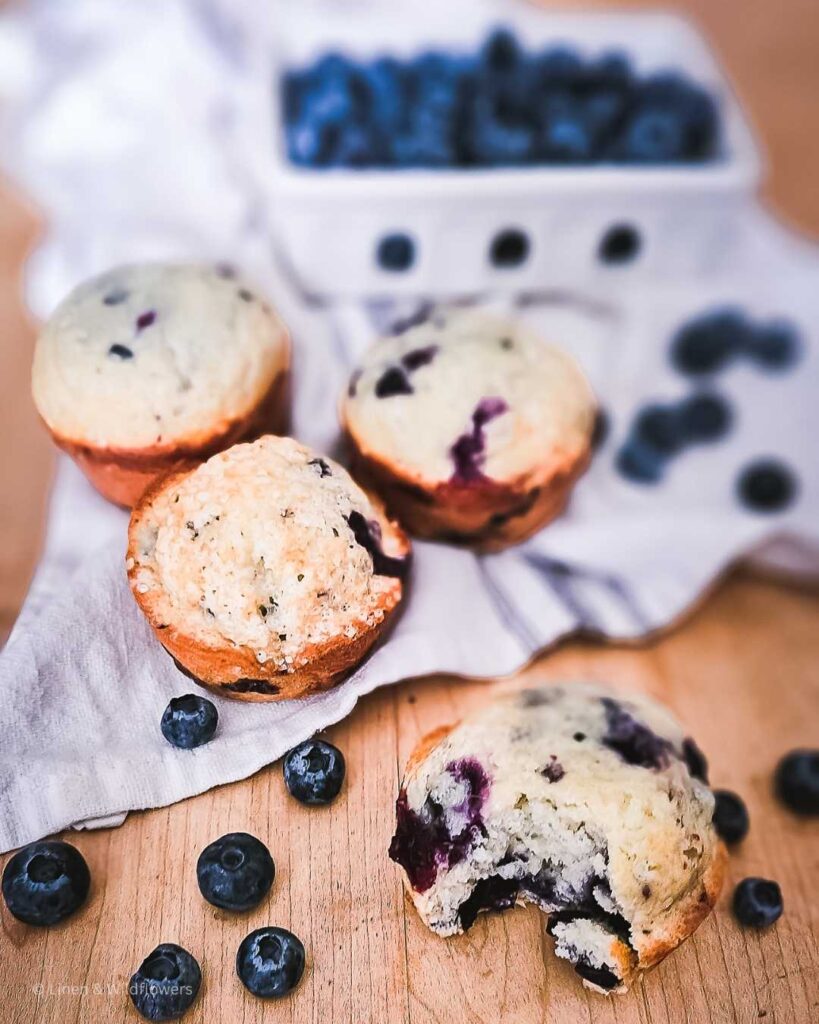 Bursting with the goodness of fresh blueberries, these muffins on a tea towel with fresh blueberries in a glass container & scattered around the freshly baked muffins. A muffin with a bite taken out.