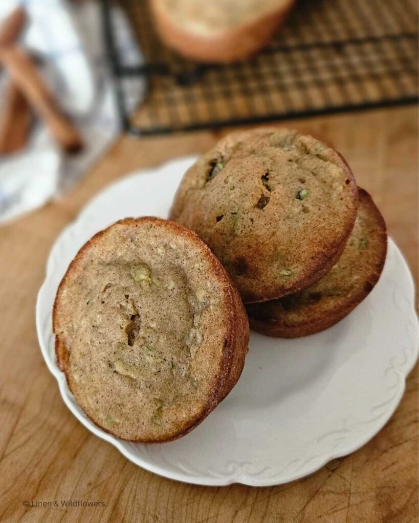 Three Zucchini Banana muffins on a white ironstone plate. Behind it, is a cooling rack for the freshly baked muffins to cool down. Next to that i a tea towel with three cinnamon stcks.