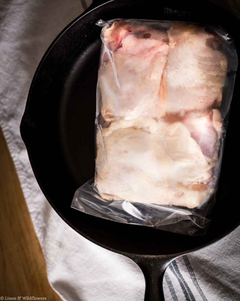 Raw chicken breasts thawing in a sealed bag inside a cast iron skillet, a simple kitchen hack for faster defrosting.