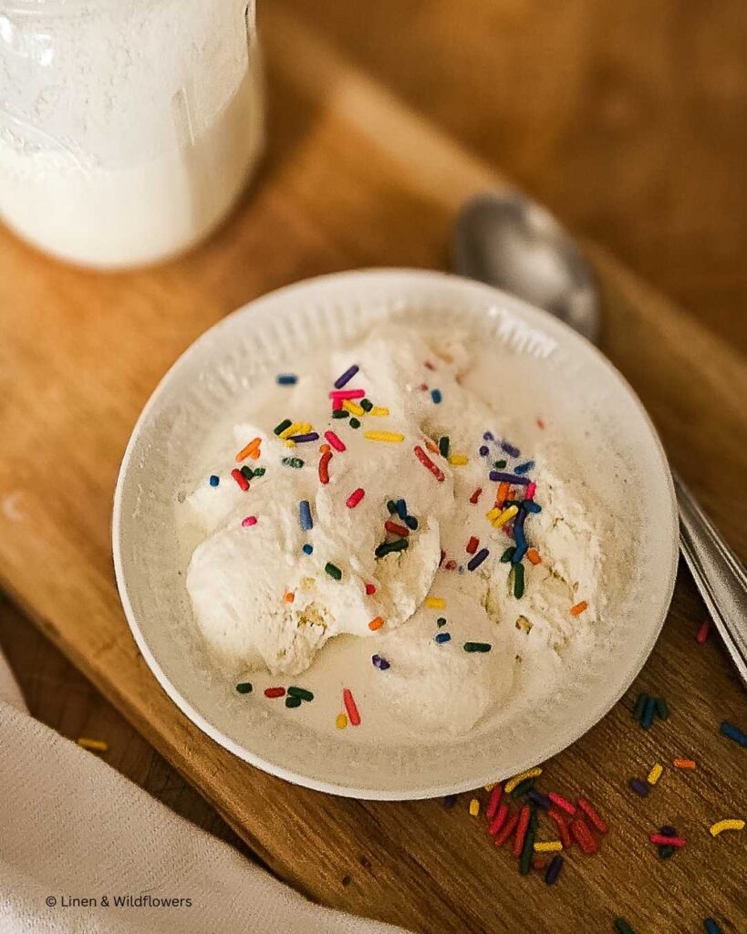 A delicious bowl of homemade vanilla ice cream with rainbow jimmies for the topping on a cutting board. Next to it is a tea towel & a mason jar with the rest of the ice cream.