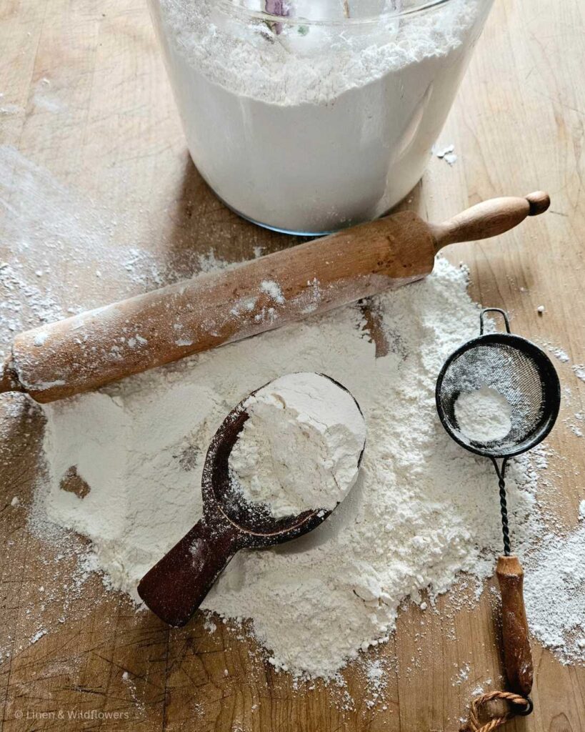 A large jar of flour next to a layer of flour on the butcher block with a rolling pin, scoop & sifter.