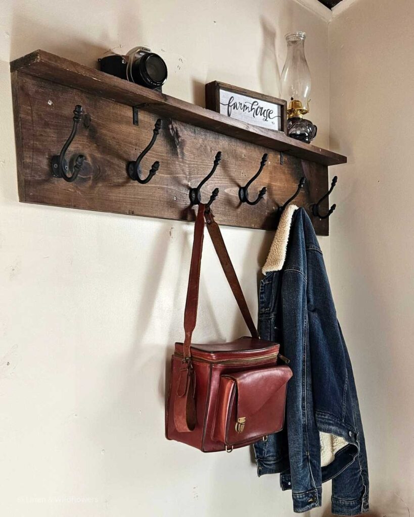 A DIY Coat rack mounted on the wall with a vintage camera bag & denim coat hanging from it. On top of the shelf is a oil lamp, SLR Canon camera & a rustic farmhouse sign.