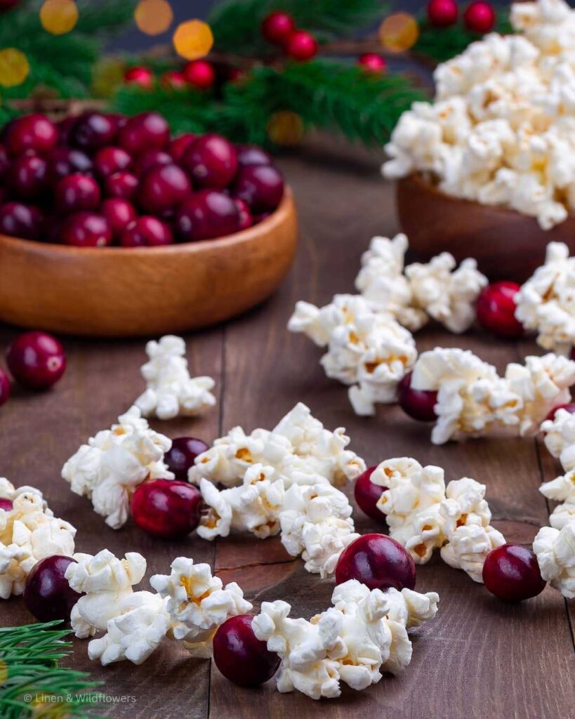 A garland with cranberries & popcorn strung next to two wood bowls each filled with cranberries & popcorn to make a homemade Christmas tree garland.