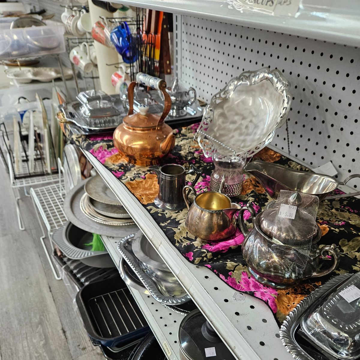 Thrift store shelves filled with an assortment of dishes & kitchenware including silver & copper serving dishes.
