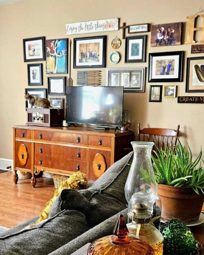 A wall gallery filled with art & family photos about a sideboard that hold a flat screen tv & a vintage stereo where a tabby kitten sitting on to look towards the living room window.