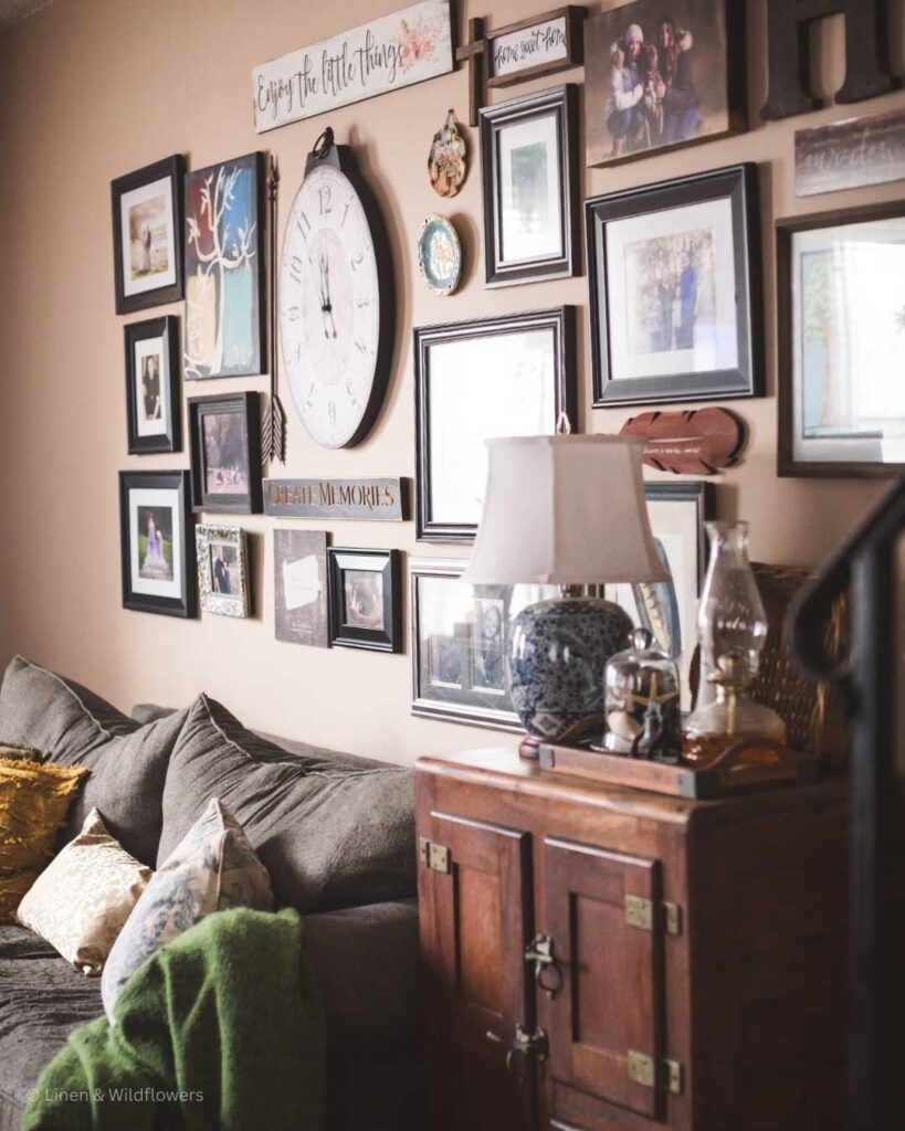 A curated wall gallery above a comfy couch with a throw pillow & blanket. Next to the couch is an antique icebox that serves as a end table that holds an oriental lamp, oil oil lamp, cloche filled with sea shells & a basket propped against the wall.