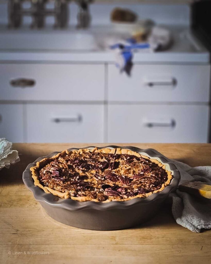  a freshly baked Old-Fashioned Southern Pecan Pie in a stone pie plate cooling on a butcher block in front of a 1950s vintage sink. Above the sink is a two tiered wall shelf with vintage mason jars filled with dry goods. On the sink drainboard is a blue & white ta towel * a scrub brush.