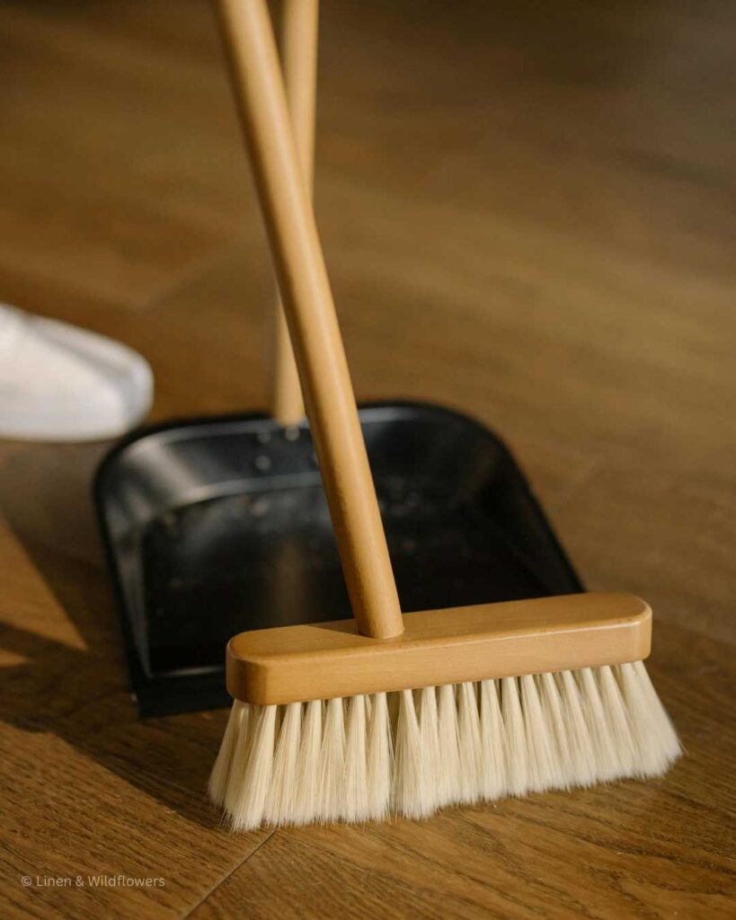Dusty sweeping the hardwood floors with wooden broom & collecting the first with a metal dust pan.