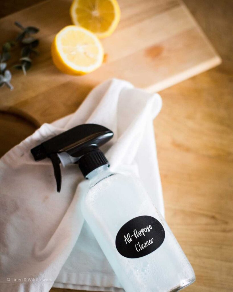 DIY All-Purpose Cleaner in a clear glass bottle on a tea towel next to a cutting board with cut lemon.