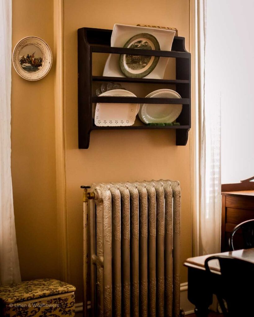 A cast iron radiator in a home built in 1885. Above the radiator is a plat rack filled with various of platters. Th to the left of the radiator is a floral sewing box.
