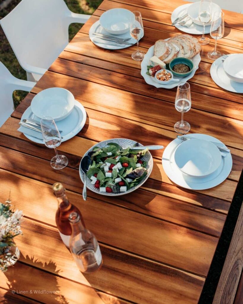 A wood picnic table set with dishes, wine glasses & platters of yummy food for guest in the backyard.