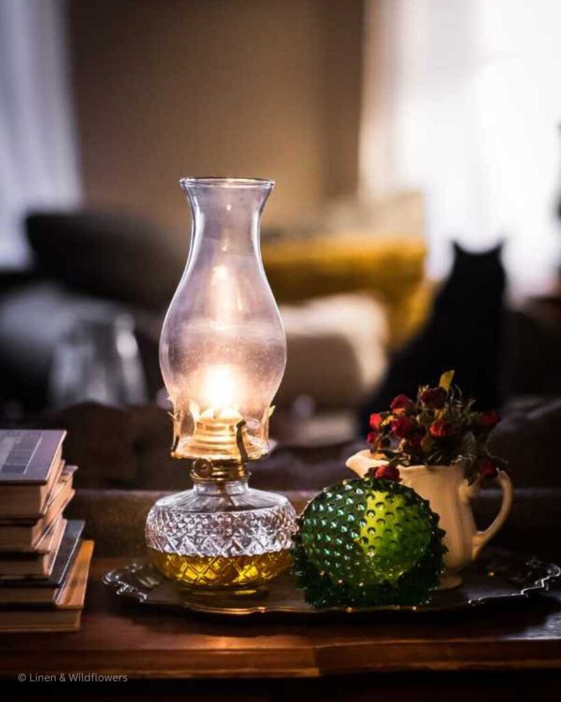 A lit vintage oil lamp on a sofa table next to a half circled green fenton dish, an ironstone creamer used to hold hold mini dried red roses on a gold plater. Next to it is a stack of home decor books.