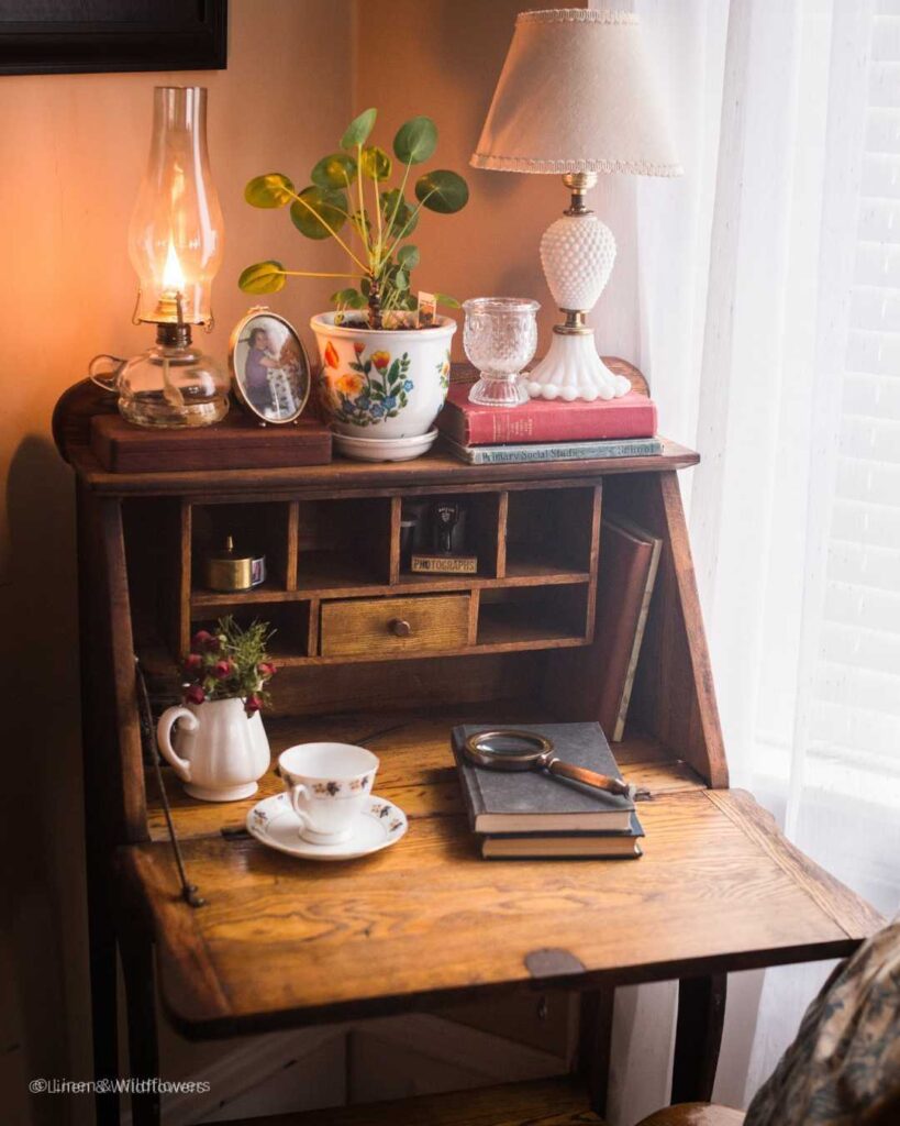 A antique secretary desk curated with vintage finds, a lit oil lamp & house plant.