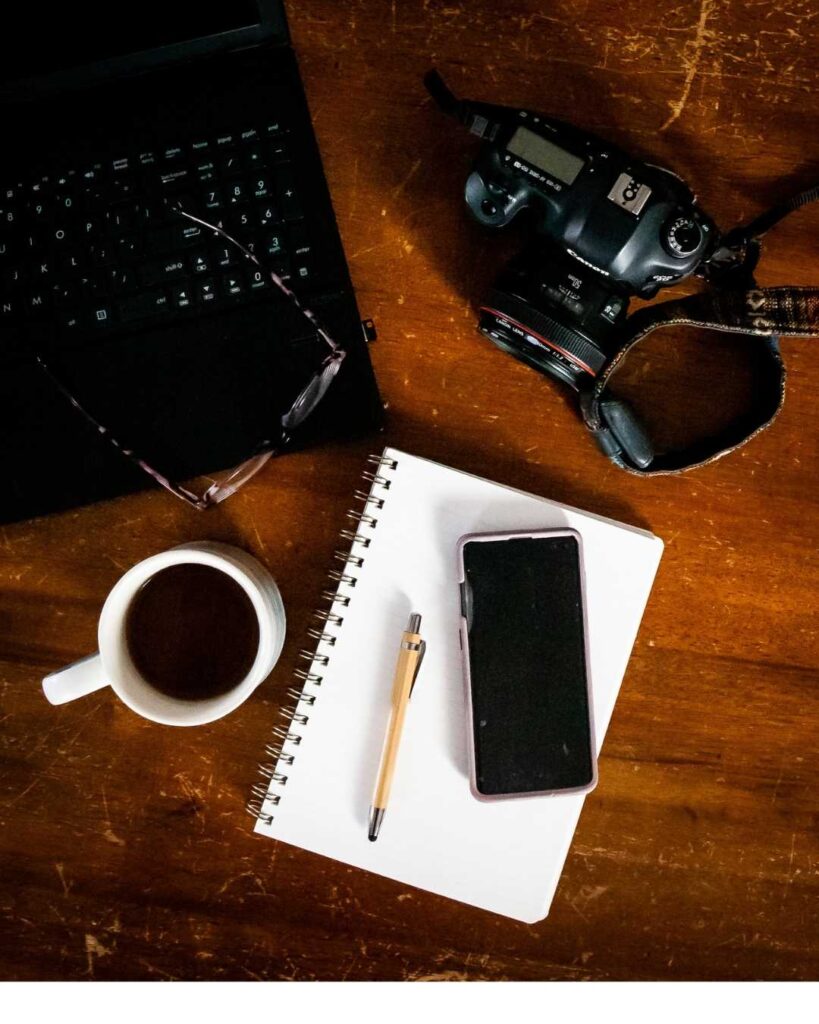 A laptop, cup of coffee, notebook with a pen & cell phone on top of it & a canon camera.