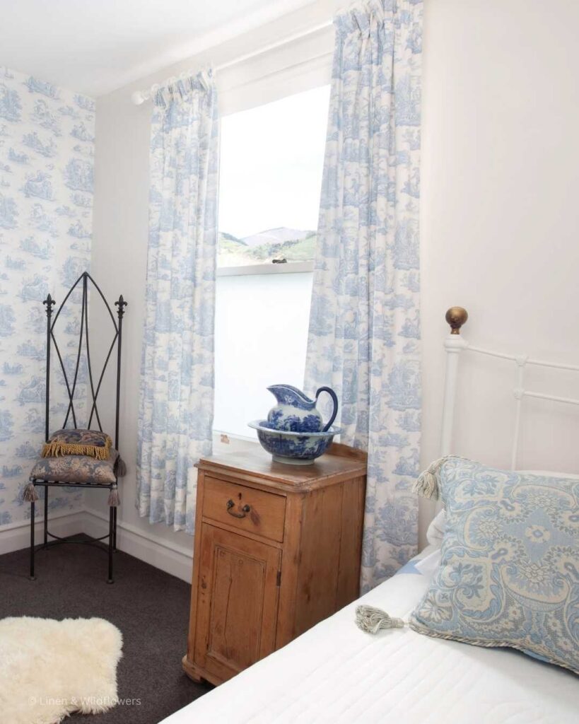 A country cottage styled bedroom. Next to a white metal bed frame is a wooden bed table with a colonial blue & white printed pitcher & bowl. On the bed is layer with a beautify printed light blue linens. In the corner of the room is a metal chair with a arched backing.