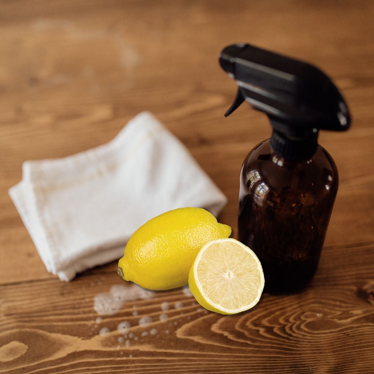Amber spray bottle, fresh lemons, and a white cloth on a wood table, showing cleaning hacks that save time and energy.