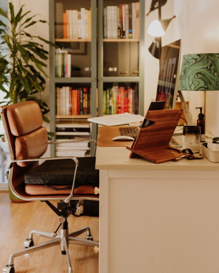 A cozy office with a leather & metal chair, a wood & metal white desk with a laptop, lamo & the necessary office supplies nneded for a home office. In the background a closed glass door book case filled with books.