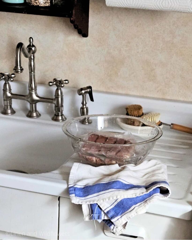 Frozen meat thawing in a clear glass bowl of water on a vintage sink.