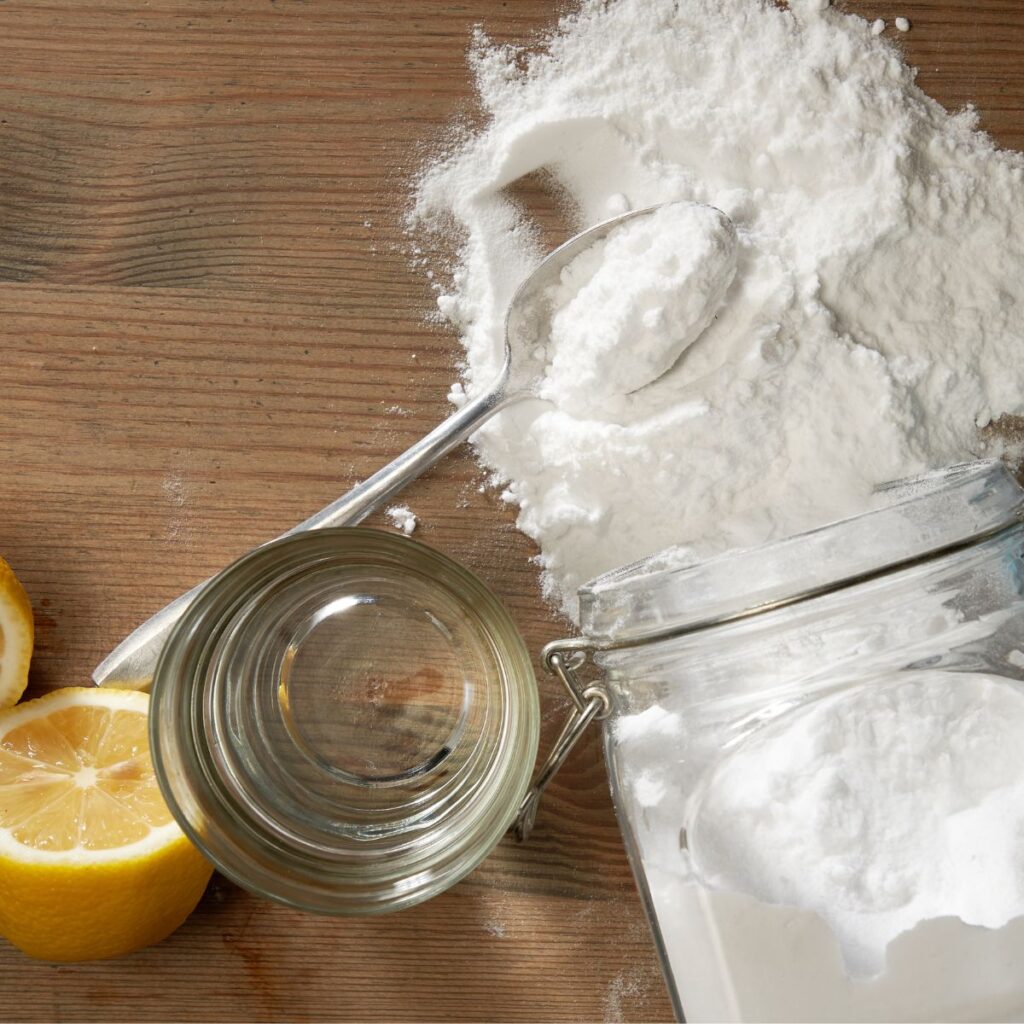 A glass of water & a jar of baking soda spilled over on a wood butcher block next to cut lemons & a silver spoonful of baking soda top of a pile of baking soda.